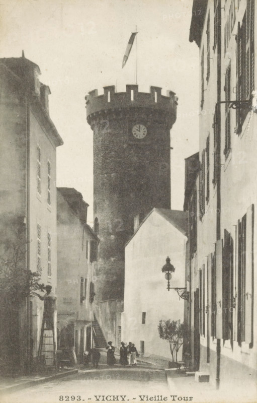 View of the Tour de l'Horloge in Vichy, Vichy.-Vieille Tour, photomechanical print, stereograph, number: '8293.', Vichy, c. 1890 - c. 1910, photographer, pasteboard, paper, collotype, photoprint, height 138 mm x width 179 mm, tower, clock tower, monumental adornment, A narrow street flanked by tall buildings leads to a large round stone tower with battlements and a flagpole extending above it, the tower showing a circular clock face; the street scene includes a few people standing and walking near the base of the tower, a set of exterior stairs on the left side, shuttered windows on the building facades, an ornate wall-mounted street lamp on the right, and a small tree or shrub near the right sidewalk.