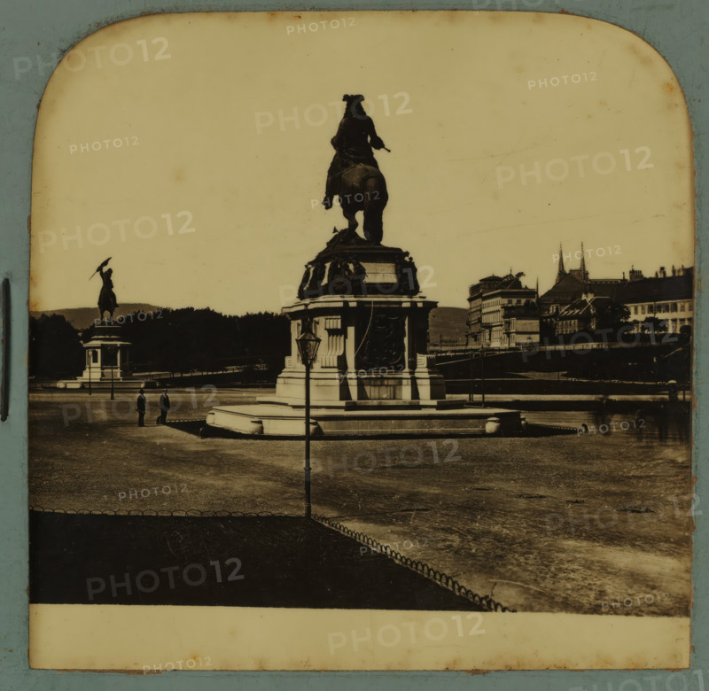 Equestrian statues of Archduke Charles and Eugene of Savoy on the Heldenplatz in Vienna, Heldenplatz in Wien, stereograph, number: '1510.', Vienna, after sculpture by Anton Dominik Fernkorn, 1900 - 1940, Anton Dominik Fernkorn, photographer, after sculpture by, Lower Austria, paper, plastic, pasteboard, organic material, cellulose nitrate film, black and white slide, height 75 mm x width 150 mm, Person, Charles V (Holy Roman Emperor and King of Spain), Eugenius of Savoy, square, circus, equestrian statue, Heldenplatz, A sepia-toned photograph showing a large equestrian statue on a stepped stone pedestal in the foreground with ornate reliefs and decorative elements, a lamppost positioned in front of the pedestal, a second equestrian statue visible further back to the left atop its own plinth, a wide open paved area surrounding the monuments with a low decorative fence edging a darker paved section, two figures standing near the base of the main pedestal, distant rows of trees and a rive...