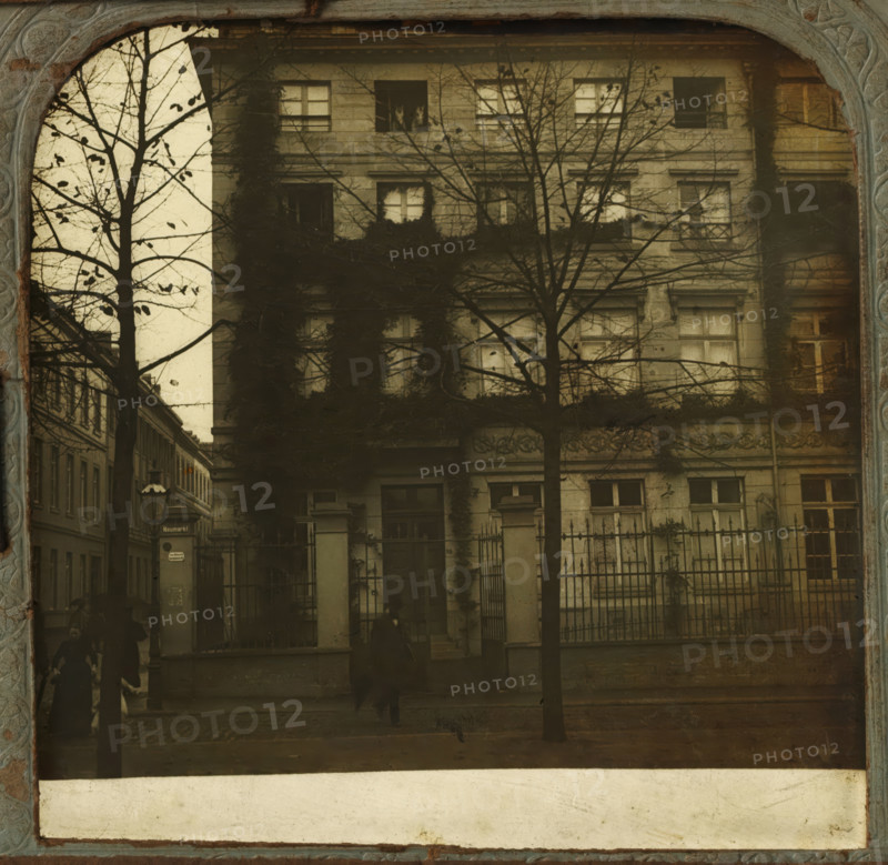 Part of the facade of the Richmodishaus in Cologne, Richmodis-Haus in Coln, stereograph, number: '575.', Cologne, 1900 - 1940, photographer, North Rhine-Westphalia, paper, plastic, pasteboard, organic material, inorganic material, cellulose nitrate film, metal, gelatin silver print, developing gelatin silver print, height 75 mm x width 150 mm, facade (of house or building), Richmodishaus, A faded sepia-toned photograph shows the facade of an older multi-story building with rectangular windows, some framed by climbing ivy and partially obscured by bare tree branches in the foreground; an iron fence with stone pillars runs along the front, and shadowy figures move along the sidewalk, their forms blurred; a narrow street and neighboring buildings flank the left side, and a streetlamp and a vertical signpost are visible near the corner.