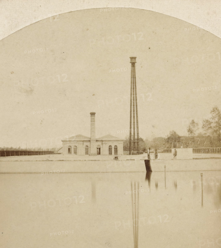 Machines of the Dune Water Supply at Leiduin, Installation for the dune water supply in Leiduin, Stereoscopic Views in the Netherlands, stereograph, initials: 'PO', inscription: 'Available at A. Jager, Water 110, in Amsterdam.', inscription: 'Netherlands', photographer: Pieter Oosterhuis, Leiduin, publisher: Andries Jager, Amsterdam, 1857 - 1865, pasteboard, paper, albumen print, height 85 mm x width 173 mm, pumping installation, pumping-engine (, polder), mineral water, A sepia-toned scene showing a small brick building with arched windows and a chimney, a tall slender metal tower beside it, a flat rooftop area in front with a few standing figures, distant trees along the horizon, and the lower portion of the image reflecting vertical shapes in a lighter expanse.
