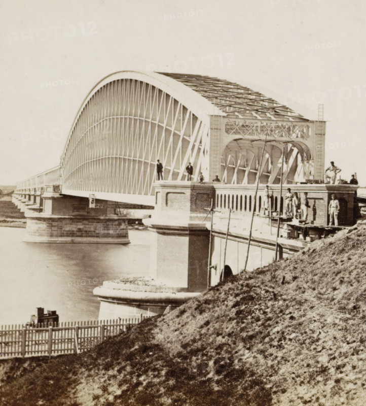 View of the Kuilenburg railway bridge near Culemborg, Bridge at Kuilenburg, stereograph, photographer: Pieter Oosterhuis, Culemborg, before 1868-09-06, pasteboard, paper, albumen print, albumendruk, height 86 mm x width 177 mm, bridge in village across river, canal, etc. - II - ideal landscapes, Kuilenburg railway bridge, A large arched metal bridge spans across water with a series of vertical and diagonal lattice supports forming the arch and deck, resting on stone piers rising from the river; workers stand and sit on the bridge structure and on the brick abutment, some near scaffolding and ladders, while wooden poles and construction materials are visible on the walkway; a grassy embankment slopes down toward a picket fence in the foreground, and calm water reflects parts of the bridge and piers.