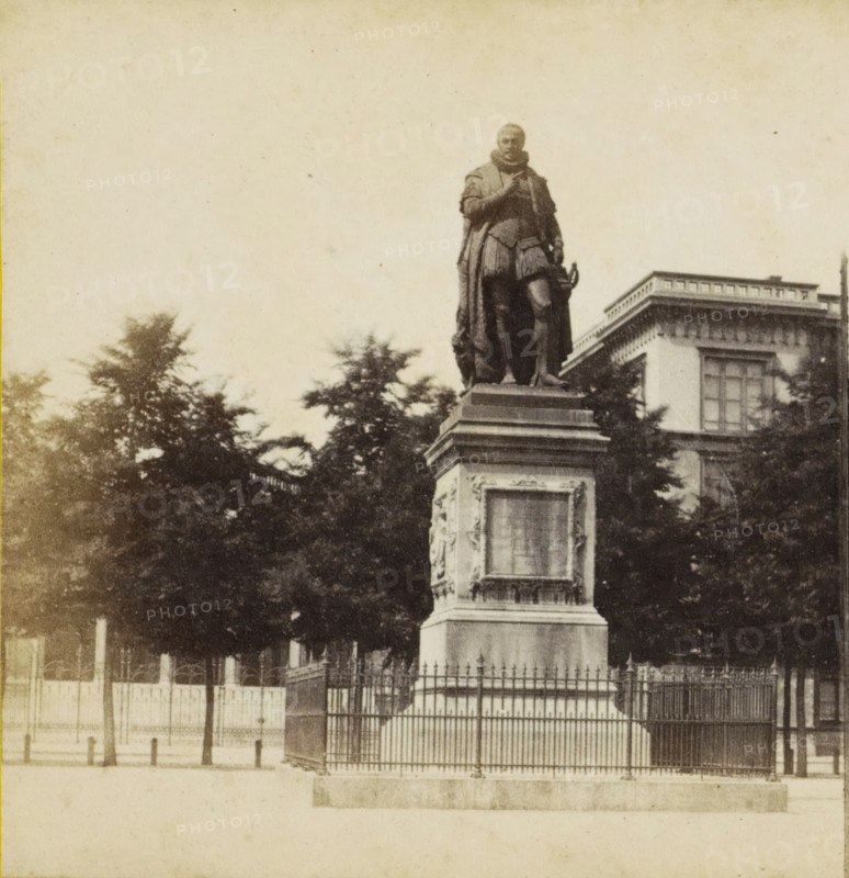 The Hague, Plein, View of the statue of William of Orange on the Plein in The Hague, stereograph, after sculpture by Louis Royer, 1873 - 1890, Louis Royer, photographer, after sculpture by, pasteboard, paper, albumen print, albumendruk, height 85 mm x width 169 mm, Person, Willem I (Prince of Orange), monument, statue, square, circus, A monochrome photograph showing a large statue of a robed figure standing on a tall, decorated stone pedestal behind an iron fence, with trees partially obscuring the lower part of the monument and a multi-story building with rectangular windows visible in the background, all set against a pale sky.