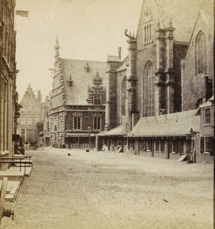 Kostershuis Haarlem, View of a street in Haarlem, stereograph, inscription: 'RBK', Haarlem, 1873 - 1890, photographer, pasteboard, paper, albumen print, height 85 mm x width 169 mm, urban housing, street, A sepia-toned street scene showing a wide cobbled road flanked by tall ornate stone buildings with pointed roofs and large arched windows, a long covered colonnade along one side with wooden posts and a sloped roof, decorative facades with sculpted details and pinnacles, a horse-drawn carriage in the distance near the buildings, benches and steps along the near sidewalk, and small bollards lining the street.