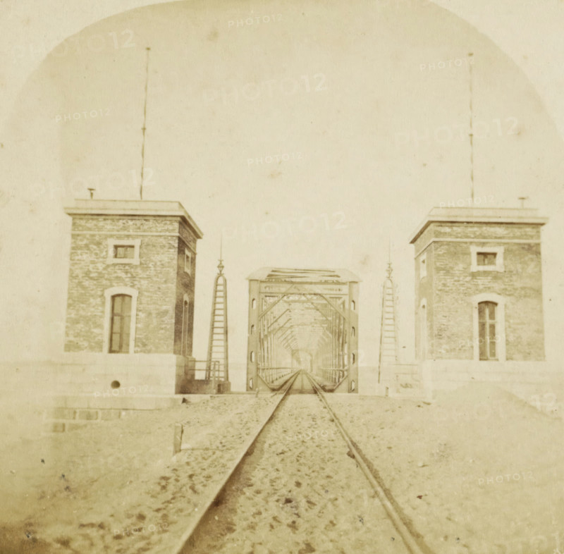 Netherlands, Great railway bridge at Moerdijk, completed 1871, View of the great railway bridge at Moerdijk, stereograph, Moerdijk, 1871 - 1872, photographer, pasteboard, paper, albumen print, height 86 mm x width 174 mm, railway, tramway, rack railway, bridge, Moerdijk railway bridge (1872), A sepia-toned scene showing a straight set of railroad tracks leading toward a metal truss bridge framed by two brick towers with rectangular windows and flat roofs, ladders and decorative finials flanking the bridge entrance, gravel and scattered debris along the track foreground, and a large pale sky occupying the upper portion of the image.