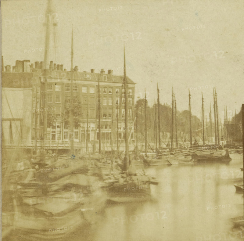 Vue prize a Rotterdam, View of the Old Harbor in Rotterdam, Hollande, stereograph, inscription: 'Rotterdam', inscription: 'Nieuwe Haven', number: 'N. 36.', photographer: Charles-Henri Plaut (possibly), Rotterdam, publisher: attributed to Alexis Gaudin et Frère, Paris, 1858, Attributed to, pasteboard, paper, albumen print, albumendruk, height 84 mm x width 167 mm, harbour, city-view in general, 'veduta', ships (in general), Oudehaven, A faded sepia view of a waterfront scene showing tall slender masts rising from moored boats in the foreground and midground, with a row of multi-story buildings behind the boats; the buildings have multiple windows and a pitched roof with chimneys, and trees are visible between and behind some masts, while the water reflects blurred shapes of the boats and masts.