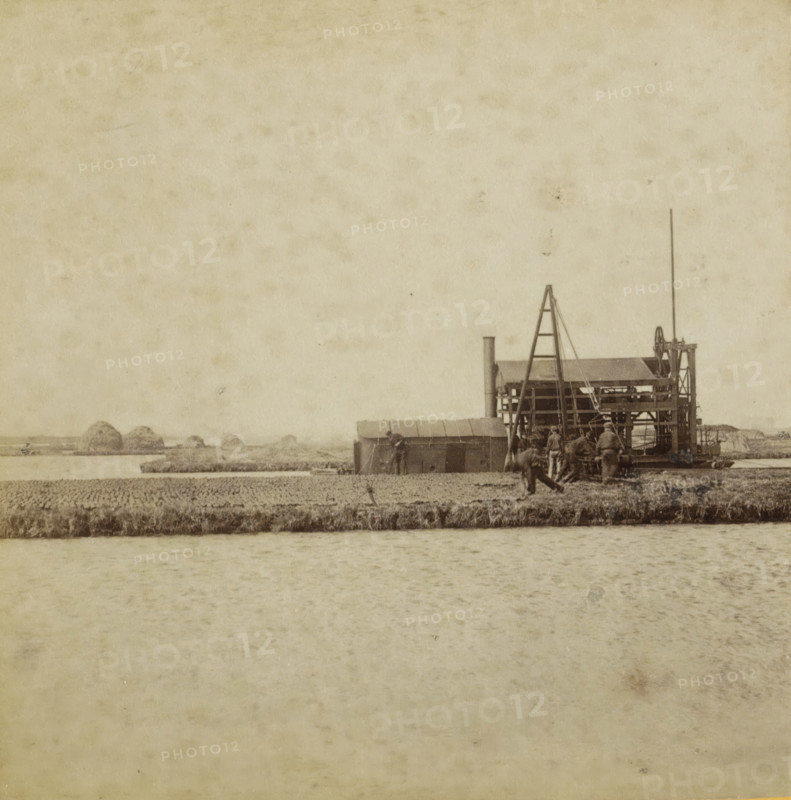 View of a state peat press machine in the low moor southeast of Amsterdam, In the low moor, Collection A, Geographical sculpture gallery, collected by MJ Broekhuijsen, headmaster of a school in Amsterdam, stereograph, inscription: SE of Amsterdam. The state peat press machine extracts the peat, presses it, and cuts peat blocks from it. See pl. 24.', number: 'A.23.', number: '23.', Amsterdam, publisher: Merkelbach & Co., compiler (person in literature): Martien Johan Broekhuijsen, 1900 - 1902, Merkelbach & Co., Martien Johan Broekhuijsen, photographer, publisher, compiler (person in literature), pasteboard, paper, albumen print, albumendruk, height 87 mm x width 176 mm, peat-cutting, world fair, world exhibition, A sepia-toned scene showing a body of water in the foreground and a narrow strip of land with grassy edges above it, behind which a working structure stands near the right edge; the structure has a tall vertical pole, a ladder-like frame, a small roofed section, visible machine...
