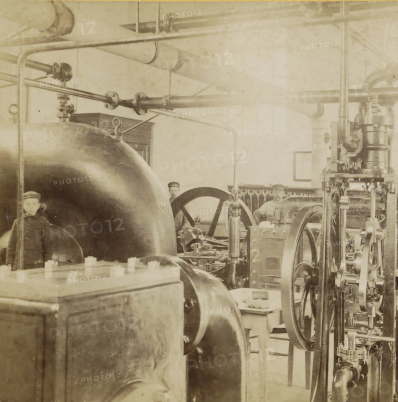 Interior of a water machine, A machine room, Geographical sculpture gallery, collected by MJ Broekhuijsen, headmaster of a school in Amsterdam, Collection E, stereograph, inscription: 'RBK', number: '5.', inscription: 'Interior of a water machine.', Netherlands, publisher: Merkelbach & Co., Amsterdam, compiler (person in literature): Martien Johan Broekhuijsen, 1900 - 1902, photographer, compiler (person in literature), pasteboard, paper, albumen print, height 86 mm x width 176 mm, machine driven by water, A sepia-toned industrial interior showing large curved metal tanks and cylindrical machinery with visible bolts and flanges, an array of pipes and horizontal rods running across the upper area, several wheels and flywheels with spokes, vertical rods and mechanical linkages, a wooden cabinet partially visible against the wall, a framed rectangle on the wall, and two partially visible figures wearing caps standing among the equipment.