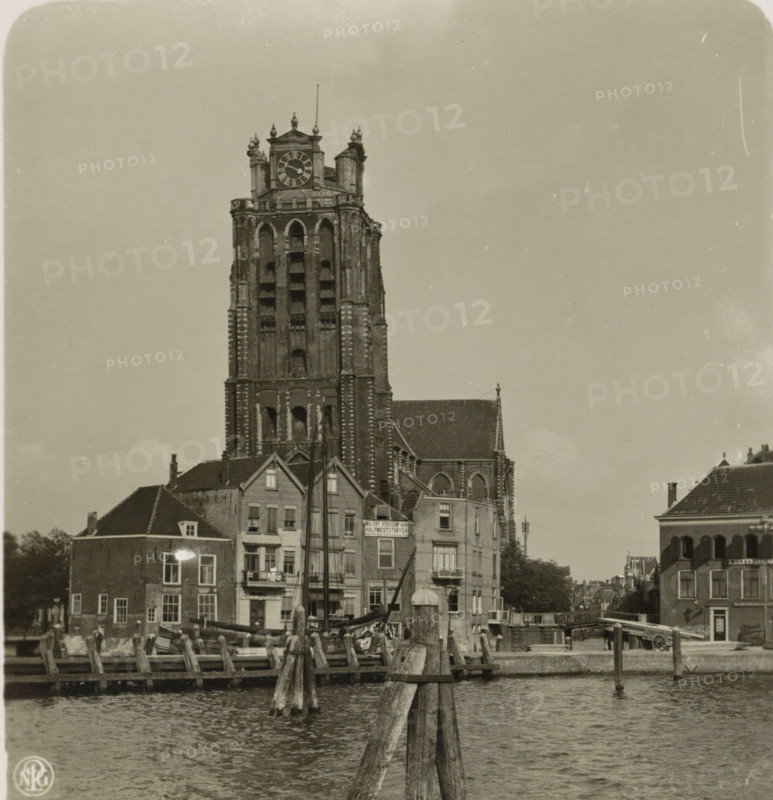 View of the Great or Our Lady Church in Dordrecht, Holland. Dordrecht. Great Church, stereograph, number: '4', Dordrecht, publisher: Neue Photographische Gesellschaft, Steglitz, 1894 - 1935, photographer, paper, gelatin silver print, developed gelatin silver print, height 88 mm x width 180 mm, parts of church exterior and annexes: tower, Great or Our Lady Church, A tall stone tower with a large round clock near its top rises above rows of adjacent buildings; the tower shows arched openings, decorative vertical ridges, and ornate pinnacles, while the lower buildings in front have pitched roofs, multiple windows, and facades with visible balconies and shutters; wooden pilings and posts extend into rippling water in the foreground, and small boats with masts are moored near the quay; a bridge and more buildings line the waterway to the right, and the sky above is clear with a few small specks and marks.