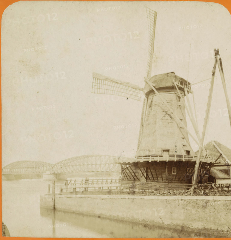 View of a mill and the railway bridge over the Oude Maas near Dordrecht, Dordrecht. Pont du chemin de fer sur la Meuse, Vues de Hollande, stereograph, number: '12', Dordrecht, publisher: J. Lévy & Cie, Paris, publisher: Léon & Lévy, 1873 - 1890, photographer, pasteboard, paper, albumen print, albumendruk, height 86 mm x width 178 mm, windmill in landscape, bridge, railway bridge, A pale, aged photograph showing the upper portion of a traditional windmill with a thatched cap and large lattice sails extending outward, the windmill's cylindrical body rising from a wooden base with a narrow balcony and supporting beams, a tall wooden tripod-like structure beside it with ropes and pulleys, a riverside foreground with a stone or concrete embankment and calm water reflecting muted tones, and an arched metal bridge spanning the river in the background under a mostly empty, lightly stained sky.