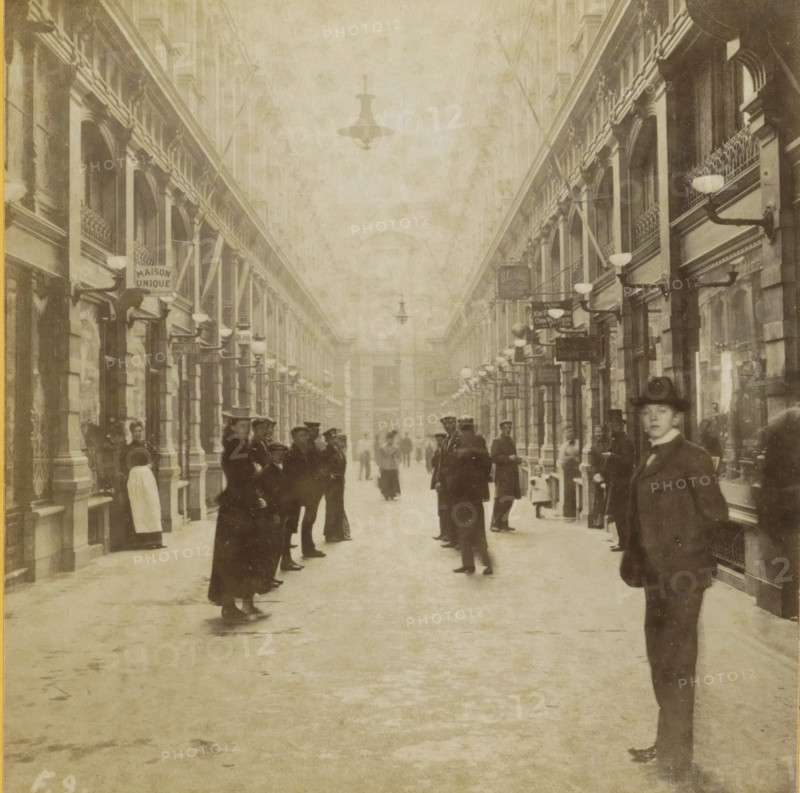 Covered shopping arcade de Passage in The Hague, Passage, Collection F, Geographical sculpture gallery, collected by MJ Broekhuijsen, headmaster of a school in Amsterdam, stereograph, inscription: 'in The Hague: covered shopping street. We see only half of it here, as the street curves at the end. Above us in the middle electric lighting.', number: '9.', number: 'F.9.', The Hague, publisher: Merkelbach & Co., Amsterdam, compiler (person in literature): Martien Johan Broekhuijsen, 1900 - 1902, photographer, compiler (person in literature), pasteboard, paper, albumen print, albumendruk, height 87 mm x width 176 mm, shop-window, show-window, arcade, architecture, A long covered passageway flanked by tall colonnaded facades with arched openings and decorative balconies lined with wall-mounted lamps, a high ceiling with hanging light fixtures, and numerous people standing and walking along the central floor; several individuals wear coats and hats, one person in the foreground on the right ...