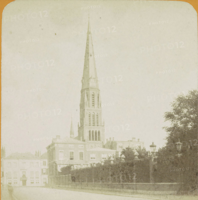La Haye. Nouvelle Eglise Catholique, View of the Sint-Jacobus de Meerkerk in The Hague, Vues de Hollande, stereograph, number: '16.', The Hague, publisher: J. Lévy & Cie, Paris, 1878 - 1890, photographer, pasteboard, paper, albumen print, albumendruk, height 87 mm x width 178 mm, parts of church exterior and annexes, Parkstraat, Sint-Jacobus de Meerkerk, A tall church steeple rises above a cluster of buildings, the steeple showing narrow vertical openings and a pointed spire with visible structural details, set against a pale sky. In front of the church are multi-story rectangular buildings with rows of windows and simple façades. A row of trees with dense foliage lines one side, partially obscuring a decorative lamppost. A low stone wall and an iron fence run along the foreground beside a paved street. The image has a faded, sepia-toned appearance and rounded corners at the top.