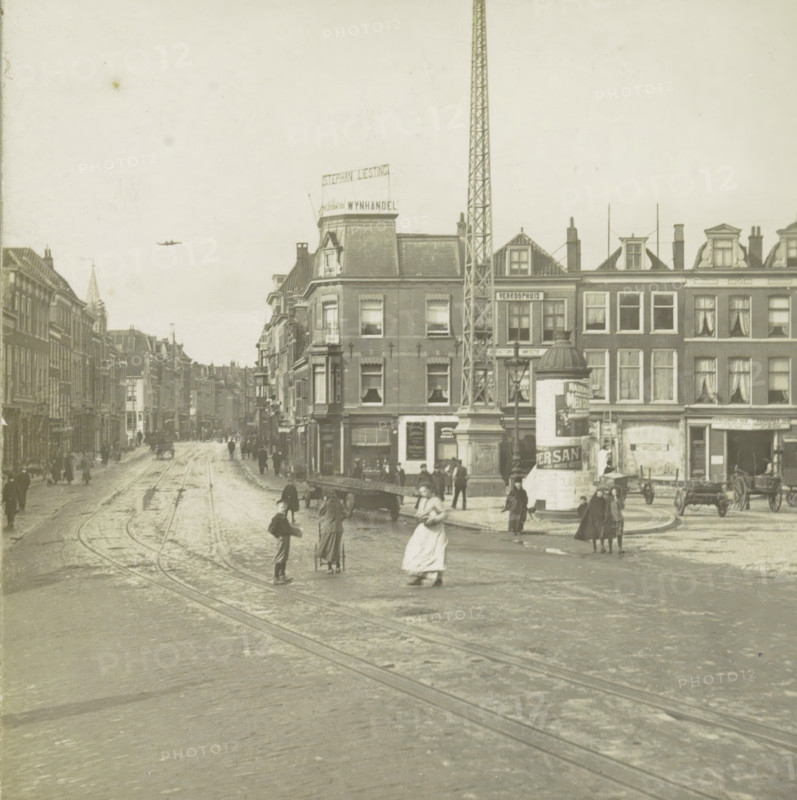 s Gravenhage.- Wagenplein, View of the Wagenplein in The Hague, stereograph, number: '112.', The Hague, 1882 - 1920, photographer, paper, pasteboard, gelatin silver print, developing gelatin silver print, height 89 mm x width 178 mm, urban housing, street, square, circus, Wagenstraat (The Hague), A wide street scene with tracks running along a paved roadway flanked by multi-story brick buildings, a tall lattice tower rising beside a corner building, an advertising column near the sidewalk, horse-drawn carts and a few figures walking or standing on the street, some people in period clothing with long skirts and coats, windows and dormers visible on the building facades, a distant row of buildings receding down the street, and a small airplane visible in the sky.