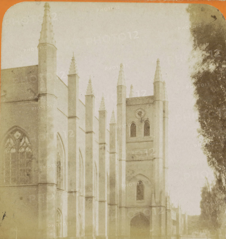 View of the Willemskerk in The Hague, La Haye. Église Guillaume, Vues de Hollande, stereograph, number: '18.', The Hague, publisher: Léon & Lévy, Paris, publisher: J. Lévy & Cie, 1873 - 1890, photographer, pasteboard, paper, albumen print, height 87 mm x width 178 mm, parts of church exterior and annexes, Willemskerk (The Hague), A faded sepia image of a large Gothic-style stone building with tall narrow buttresses topped by pointed pinnacles, arched windows with tracery, a tower section with louvered openings and a circular clock face, and a dark arched doorway at ground level, with a large dark tree partially visible along the right edge.