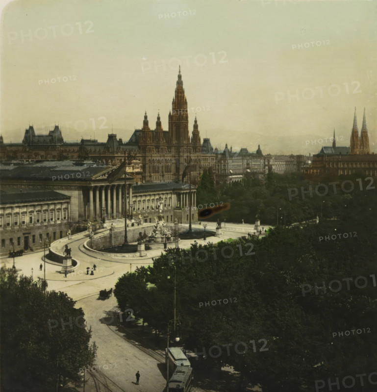Burgring with the Austrian Parliament and Viennese Town Hall in the background, Vienna, Wien. Burgring mit Parlament, stereograph, inscription: 'Oesterreich. Vienna. Burgring mit Parlament., Austria. Vienna 'Burgring' with Parliament., Autriche. Vienne. 'Burgring' et Parliament., Austria. Viena. 'Burgring' y Parlamento.', monogram: 'BW', number: '17.', photographer: BW, 1898 - 1935, Lower Austria, paper, baryta paper, gelatin silver print, hand coloring, developing gelatin silver print, height 88 mm x width 169 mm, city-view in general, 'veduta', A wide aerial view of a cityscape featuring a large ornate building with tall pointed towers and intricate facades centered in the midground, flanked by other classical buildings with columns and decorative roofs, a curved plaza with fountains and statues in front of the buildings, tree-lined streets and dense dark foliage occupying the lower half of the image, small figures and horse-drawn carriages on the plaza and streets, a tram on tracks ...