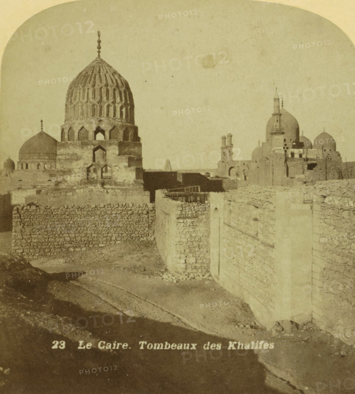 Le Caire. Tombeaux des Khalifes, Graven van de Mammelukse kalifen te Caïro, stereograph, inscription: 'C', number: '23', number: '25343', photographer: JG Beers, Caïro (city), publisher: Nederlandsche Stereoscoop Maatschappij, Amsterdam, 1873 - 1890, Ottoman Empire, North Africa, Cairo (city), pasteboard, paper, albumen print, albumendruk, height 88 mm x width 179 mm, slavery, serfs and enslaved persons, grave, tomb, A sepia-toned photograph shows a skyline of domed stone buildings and mausoleums behind a high stone wall; a large ornate dome with patterned vertical ridges and arched openings at its base dominates left of center, flanked by smaller domes and towers with finials and small windows; the foreground features a rough stone embankment and a curved pathway or dry channel leading alongside the textured masonry wall, with scattered rubble and a small stepped section; in the distance more domed structures and parts of ruined walls rise against a pale sky.