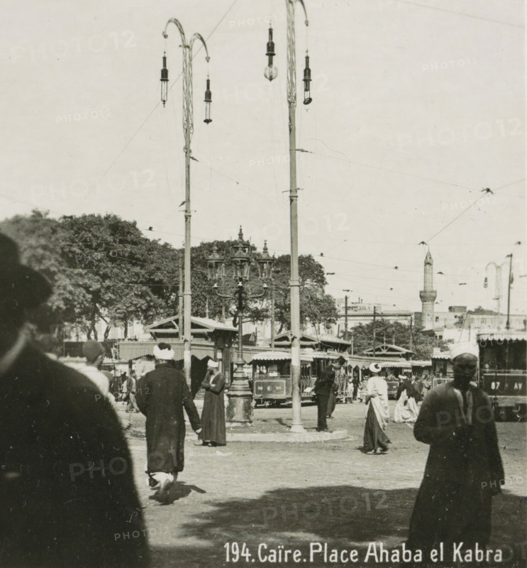 Caire. Place Ahaba el Kabra, View of a square in Cairo, stereograph, number: '194.', inscription: 'These stereo-photographs are being offered for sale and sold wholesale and retail by the ........., ', inscription: 'Submitted.', monogram, address: 'Parklaan 49, HAARLEM, (Holland)', photographer: Richard Sochor, Cairo (city), publisher: De Wereld-Tourist, Haarlem, 1903, Ottoman Empire, North Africa, paper, baryta paper, gelatin silver print, gelatin silver developing print, height 88 mm x width 179 mm, square, circus, A crowded outdoor scene with tall ornate lamp posts and hanging streetlights above a plaza, trees and a minaret visible in the background, a central decorative multi-lantern street lamp, market stalls or small structures with awnings, a tram or streetcar partially visible behind the lamp, numerous people in period clothing and head coverings walking or standing in the foreground, overhead wires crossing the sky.