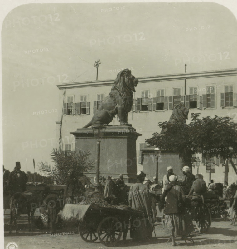Entrance of Qasr al-Nil Bridge, Cairo, Cairo. Die Brücke von Kasr el Nil, stereograph, number: '6', Qasr al-Nil Bridge, publisher: Neue Photographische Gesellschaft, Steglitz, 1931 - 1935, photographer, Ottoman Empire, North Africa, paper, baryta paper, gelatin silver print, developing gelatin silver print, height 90 mm x width 180 mm, sculpture, bridge, A large stone lion statue sits atop a rectangular pedestal in front of a multi-windowed building, another lion statue is partially visible behind foliage, a lamp post is mounted on the pedestal, people wearing headwraps and cloaks gather and stand around carts and animals, a wooden cart with large spoked wheels and a blanket-draped load is in the foreground, shutters cover many windows of the building, potted palms and trees appear near the base of the pedestal, and the sky fills the upper portion of the image.