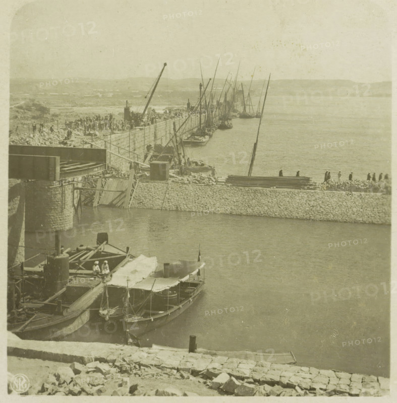 View of the Lower Aswan Dam, Egypt, Nilstaudamm bei Assuan, Upper Egypt, stereograph, number: '39.', number: 'S 20246', Lower Aswan Dam, publisher: Neue Photographische Gesellschaft, Steglitz, 1909 - 1935, photographer, North Africa, Aswan, Lage Aswandam, paper, baryta paper, daylight gelatin silver print, height 88 mm x width 180 mm, barrage, A sepia-toned harbor scene shows a stone quay curving along calm water with multiple small boats moored alongside, several with canvas awnings and visible rigging and masts rising vertically and at angles; a stone wall and piled rocks form the quay edge with a group of people standing and walking along it, and further back more people and construction materials are visible on the shore; cranes and wooden beams extend over the quay and boats, and a larger structure with stone piers projects into the water at the left; distant low hills form the background under a pale sky.