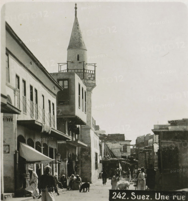 Suez. Une rue, View of a street in Suez, stereograph, address: 'Parklaan 49, HAARLEM, (Holland)', inscription: 'Déposée.', monogram, inscription: 'These stereo photographs are marketed and sold wholesale and retail by the ........., ', number: '242.', photographer: Richard Sochor, Suez, publisher: De Wereld-Tourist, Haarlem, 1903, North Africa, paper, baryta paper, gelatin silver print, developing gelatin silver print, height 88 mm x width 179 mm, street, A narrow street scene with multi-story buildings lining the left side, featuring balconies, awnings, and shuttered windows, and a tower with a conical roof and a small balcony near the top rising above the rooftops; people in traditional clothing walk and stand along the street, some clustered near shopfronts, and a few animals are visible on the ground; awnings and street lamps project from the buildings, and the overall image has a monochrome, aged appearance.