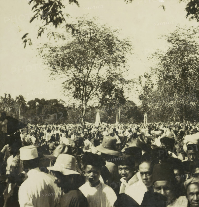 Crowd of people in a square, possibly on Borneo, Indonesia, stereograph, 1900 - 1940, photographer, paper, pasteboard, baryta paper, gelatin silver print, height 90 mm x width 178 mm, crowd, mob, A crowded outdoor scene with many people gathered closely together in the foreground and extending into the middle ground, several wearing hats of different shapes and light-colored clothing; a tall tree with sparse foliage rises behind the crowd, flanked by other trees and dense vegetation, and an obelisk-like monument is visible among the people in the distance; the sky above is light and mostly clear with a few overhanging branches visible at the top edges.