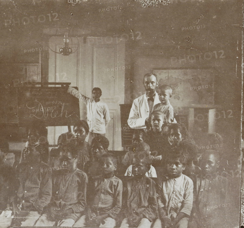 School class with teacher in Manokwari, Indonesia, Dorei. School, Collection D, Geographical gallery of images, collected by MJ Broekhuijsen, headmaster of a school in Amsterdam, stereograph, number: '17.', inscription: '(See pl. 16.) In the background by the blackboard stands the guru, (native teacher, ) who has been appointed by the missionaries in the school also founded by them., ', Manokwari, publisher: Merkelbach & Co., Amsterdam, compiler (person in literature): Martien Johan Broekhuijsen, 1900 - 1902, photographer, compiler (person in literature), paper, pasteboard, baryta paper, daylight gelatin silver print, height 89 mm x width 177 mm, elementary school, class, form, A faded sepia photograph showing a group of children seated in rows in front of a man who stands holding a smaller child, with another child standing beside a chalkboard and a tall door behind them; the room includes a hanging light fixture near the ceiling and a framed map or picture on the wall, and the image ...