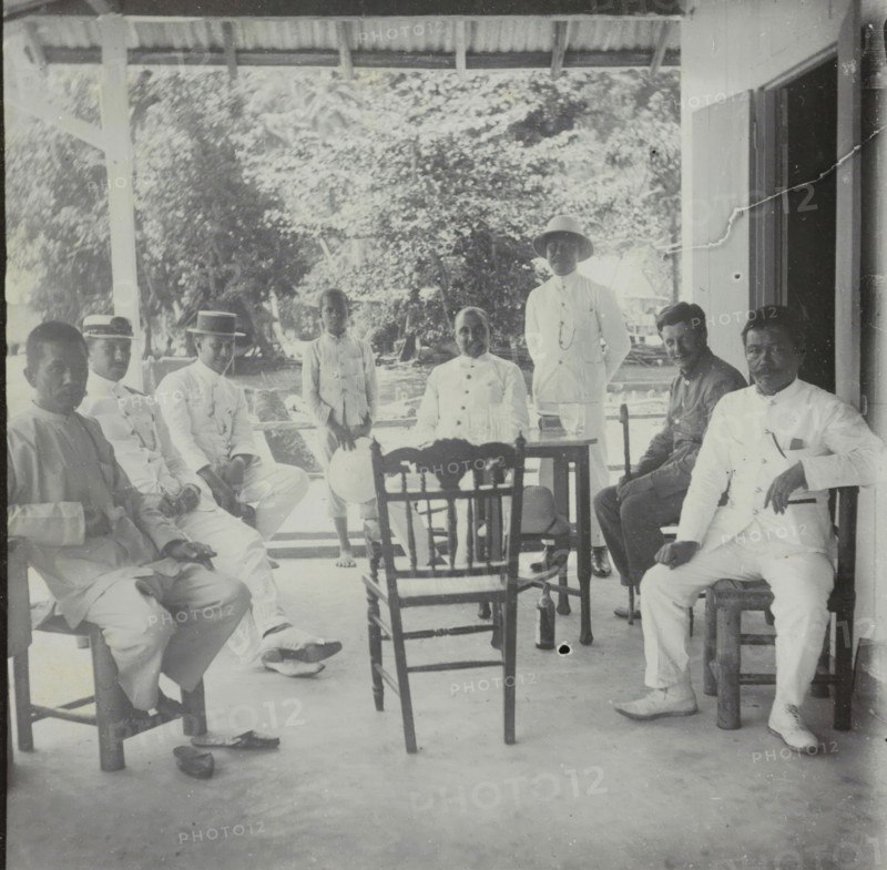 Manoekwari, Seven men and a child on a veranda in Manokwari, West Papua, stereograph, Manokwari, 1900 - 1940, photographer, paper, pasteboard, baryta paper, gelatin silver print, developing gelatin silver print, height 87 mm x width 178 mm, loggia, verandah, porch, A group of men and a boy sit and stand on a covered porch with wooden posts and a corrugated roof, surrounded by leafy trees in the background; several men wear light-colored clothing and some wear hats including a boater and a pith helmet, while others wear darker shirts; wooden chairs and a small table with glassware sit in the center of the scene, one hat rests on a chair and another on the floor, and shoes are visible near the chairs; the porch floor shows scuff marks and a small dark spot, and a doorway or open wall is visible at the right edge of the image.
