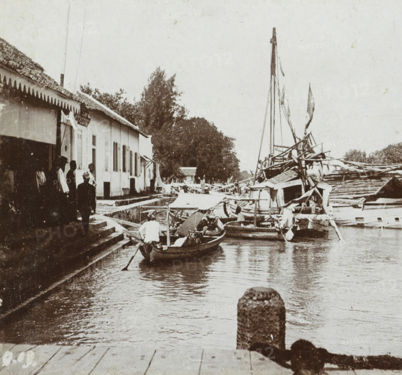 Harbor and toll office in Surabaya, Soerabaja. Small tree, Geographical gallery of images, collected by MJ Broekhuijsen, headmaster of a school in Amsterdam, Collection D, stereograph, number: 'D.19.', inscription: 'De Boom is the landing place for small vessels (such as praus) coming from the large boats to the toll office, ', Surabaya, publisher: Merkelbach & Co., Amsterdam, compiler (person in literature): Martien Johan Broekhuijsen, 1900 - 1902, photographer, compiler (person in literature), Java, East Java, paper, pasteboard, baryta paper, daylight collodion silver print, height 88 mm x width 177 mm, harbour, toll gate, A riverside scene showing a row of low buildings with tiled roofs and awnings along a stepped stone quay, several people standing on the steps and by the building, a small wooden boat with a person rowing in the water near the quay, larger boats with masts and rigging moored along the opposite side, tarpaulins and canopies covering parts of the boats, trees and fol...
