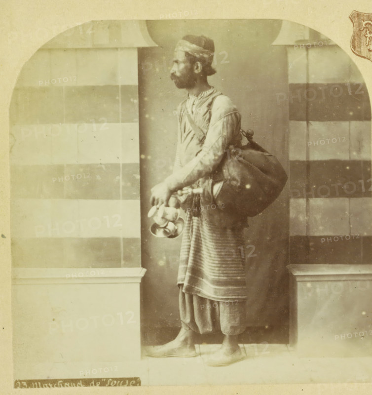 Koopman te Sousse, Tunisia, Marchand de 'Souse', Palestine et Syrie, Palestine and Syria, stereograph, number: '23.', photographer: F & E, Sousse, Paris (possibly), 1879 - 1910, North Africa, pasteboard, paper, albumen print, height 87 mm x width 173 mm, merchant, salesman, A sepia-toned photograph of a man standing in profile, wearing a headwrap and a long sleeved tunic with a striped lower garment, a large bag slung over his shoulder, and holding a cluster of round objects and shallow bowls in one hand, positioned between two architectural pillars with a checkered wall pattern behind him and wearing pointed shoes while standing on a tiled floor.