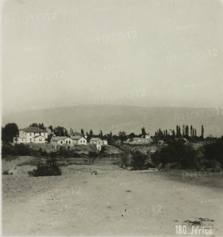 View of Jericho, Jérico, stereograph, address: 'Parklaan 49, HAARLEM, (Holland)', inscription: 'These stereo-photos are being brought into trade and sold wholesale and retail by the ........., ', monogram, inscription: 'Submitted.', number: '180.', photographer: Richard Sochor, Jericho, publisher: De Wereld-Tourist, Haarlem, 1903, Palestine, West Bank, paper, baryta paper, gelatin silver print, developed gelatin silver print, height 88 mm x width 179 mm, city-view in general, 'veduta', A monochrome photograph showing a wide sandy or gravelly foreground leading to a slightly elevated area with scattered low shrubs and vegetation, behind which a row of small buildings with pitched roofs and several larger multi-story houses sit among tall, narrow trees and other foliage, with a distant low ridge or hill visible across the middle of the image and a pale, textured sky occupying the upper portion.