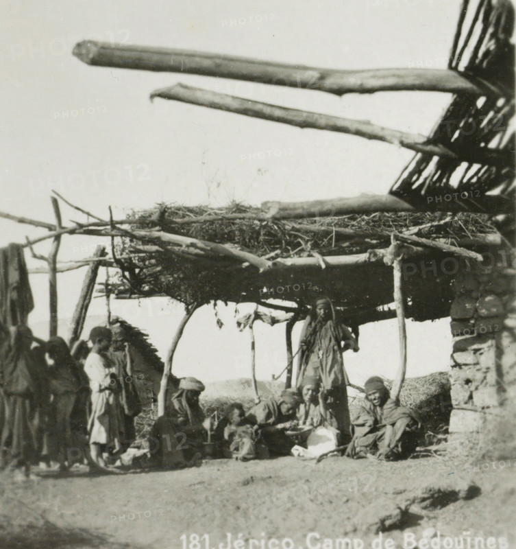 Bedouin camp in Jericho, Jérico. Camp of the Bedouins, stereograph, number: '181.', address: 'Parklaan 49, HAARLEM, (Holland)', monogram, inscription: 'These stereo photographs are being brought into trade and sold wholesale and retail by the ........., ', inscription: 'Submitted.', photographer: Richard Sochor, Jericho, publisher: De Wereld-Tourist, Haarlem, 1903, Palestine, West Bank, paper, baryta paper, gelatin silver print, developing gelatin silver print, height 86 mm x width 180 mm, tent-camp, wandering communities: nomads, A group of people wearing layered clothing and head coverings are gathered beneath a simple shelter made of rough wooden poles and a woven roof, with several large horizontal logs projecting outward above; some individuals are standing at the shelter's edge while others sit or crouch on the ground and on low stone structures, and portions of a stacked stone wall are visible at one side.