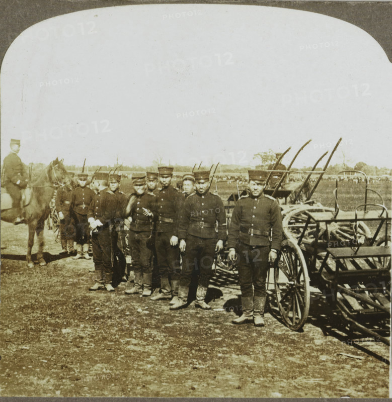 Artillery Wagons of First Division, Seoul, Korea, Korean soldiers for weaponry, stereograph, copyright notice: 'Copyright 1904, by George W. Griffith.', number: '1.', publisher: Griffith & Griffith, Philadelphia, 1904, photographer, North America, paper, pasteboard, baryta paper, gelatin silver print, developing gelatin silver print, height 89 mm x width 178 mm, the soldier, the soldier's life, (military) equipment and supply, A group of uniformed men stand in a line outdoors in front of horse-drawn equipment, each wearing dark jackets, caps, belts, and boots, with some holding items at their sides; a mounted man on a horse appears at one end of the line, several wheels and shafts of carts and equipment are visible behind and beside the men, and an open field with distant trees fills the background under a pale sky.