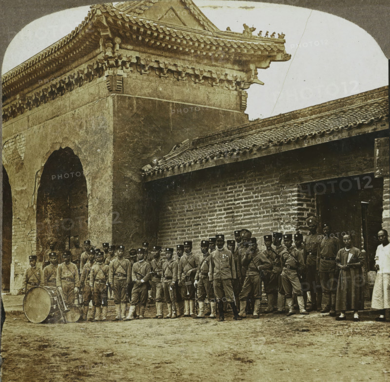 Orchestra at Seoul, Korea, A Regimental Band at Seoul, stereograph, copyright notice: 'Copyright 1904, by George W. Griffith.', number: '28.', Seoul, publisher: Griffith & Griffith, Philadelphia, 1904, photographer, North America, paper, pasteboard, baryta paper, gelatin silver print, developing gelatin silver print, height 89 mm x width 178 mm, brass band, street band, A group of uniformed men stand in formation in front of a large stone and brick building with ornate tiled roofs, decorative eaves, and arched openings; some men hold musical instruments including drums and brass instruments, several wear hats with round emblems on the fronts, and a few individuals at the edges wear long robes; the foreground shows a worn dirt ground while the upper portion of the image reveals the building's carved roofline and patterned roof tiles against a pale sky.