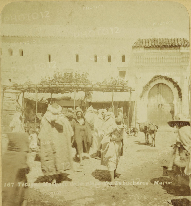 Tetuan. Mosque de la place des Babucheros. Maroc, Passers-by on the Babucheros Square in Tétouan, stereograph, number: '264', number: '167', Tétouan, publisher: American Stereoscoop Platen Gezelschap Kunst Vermaak en Onderzoek, Netherlands, 1879 - 1910, American Stereoscoop Platen Gezelschap Kunst Vermaak en Organisatie, photographer, North Africa, pasteboard, paper, albumen print, albumendruk, height 88 mm x width 178 mm, square, circus, A faded street scene showing a group of people clustered in front of a light-colored wall with a row of small arched openings and a tiled roof section at one end, some figures wearing long robes and wrapped head coverings and a few faces visible among them, a wooden canopy or stall structure with branches and foliage overhead behind the crowd, an arched doorway set into the wall decorated with a garland and secured by a horizontal bar, a load-bearing animal standing near the doorway with packs on its back, and a ground surface showing shadows and unev...