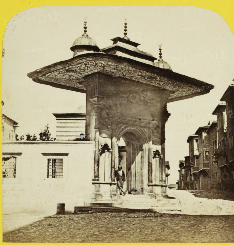 Une porte de Ste Sophie Constantinople, Gate of Topkapi Palace, Istanbul, Turquie au Stéréoscope, stereograph, number: '308', photographer: Charles Gaudin, Topkapi Palace, 1861 - 1870, pasteboard, paper, albumen print, height 84 mm x width 174 mm, gate, entrance, Constantinople, Ottoman Empire, A stone gateway with ornate carved columns and a wide decorative overhanging canopy topped by small domes and finials stands at the edge of a cobbled street, a person leaning against one of the columns beneath the archway, a low wall with barred windows adjacent to the gateway, a row of multi-story buildings with projecting upper floors lining the street to the right, steps leading up to the entrance and a worn street surface in the foreground.