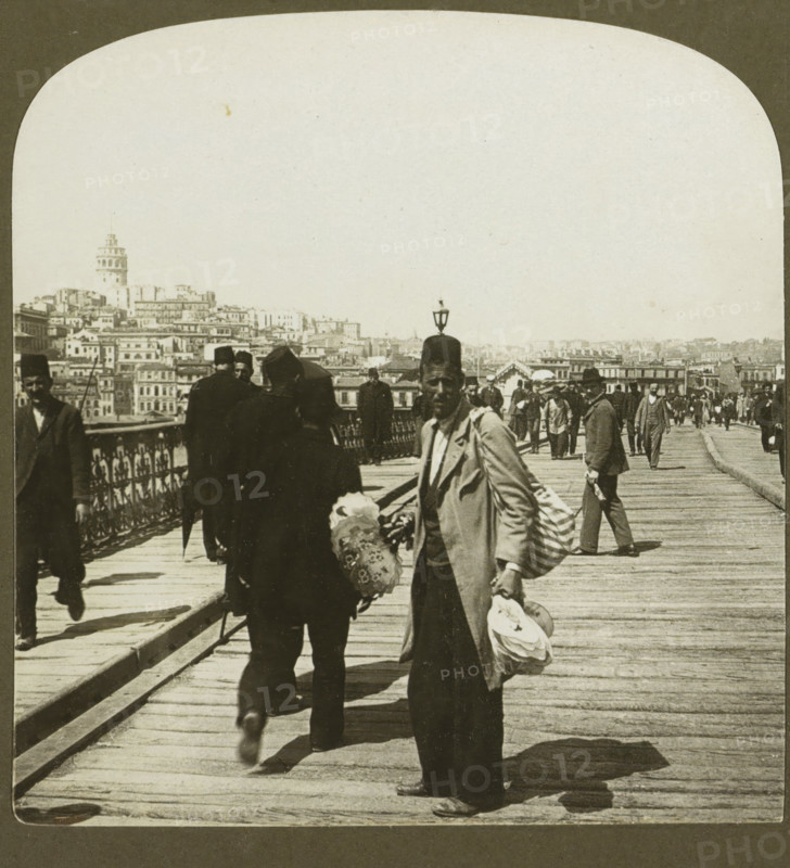 Galata, the Foreign part of Constantinople, from the Outer Bridge, Fan seller on the Galata Bridge in Istanbul, stereograph, copyright notice: 'Copyright 1901 by HC White Co.', number: '3', number: '4309.', inscription: 'The ' PERFEC' STEREOGRAPH. (Trade Mark.), Patented April 14, 1903. Other Patents Pending.', Galatabrug, publisher: HC White Co., New York (city) (possibly), 1901, photographer, Istanbul, North America, paper, pasteboard, baryta paper, gelatin silver print, developing gelatin silver print, height 88 mm x width 177 mm, pontoon bridge, Galata Bridge, A sepia-toned photograph of a wooden pedestrian bridge crowded with people, showing a foreground man facing the camera wearing a long coat and a hat, holding stacks of round objects and a patterned umbrella, with another man walking away beside him wearing dark clothing and a fez, ornate metal railings along the left side of the bridge, a row of people walking in both directions further along the bridge, a distant cityscape o...