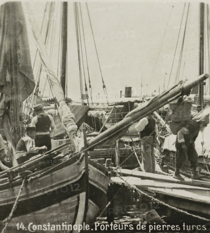 Turkish sailors in a port in Istanbul, Porteurs de pierres turcs, Constantinople, stereograph, monogram, inscription: 'These stereo photographs are marketed and sold wholesale and retail by the ........., ', number: '14.', inscription: 'Déposée.', address: 'Parklaan 49, HAARLEM, (Holland)', Istanbul, publisher: De Wereld-Tourist, Haarlem, 1903, photographer, paper, baryta paper, gelatin silver print, developing gelatin silver print, height 88 mm x width 180 mm, harbour, A crowded scene on a dock with several wooden boats and visible masts, spars, and rigging filling the frame, cloth sails and tarps draped over parts of the vessels, ropes and lines running between hulls and down to the water, a few men on and between the boats wearing vests and hats, a close-up of a boat bow and gunwale in the foreground, wooden planks and small gangways between the boats, a distant funnel and additional masts visible on the horizon, and a textured sky above.