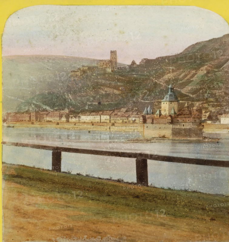 View of Kaub and the Burgen Gutenfels en Pfalzgravenstein, Germany, Pfalz, Caub et ruines du Gutenfels, Bords du Rhin, Allemagne, stereograph, inscription: 'Déposé.', number: 'N. 470', photographer: Hippolyte Jouvin, Kaub, 1860 - 1870, Rhineland-Palatinate, pasteboard, paper, albumen print, height 86 mm x width 177 mm, landscape with ruins, city-view, and landscape with man-made constructions, Burg Gutenfels, Burg PfalzGrafstein, A landscape scene showing a river in the middle ground with a long bridge crossing it supported by dark vertical piers, a grassy foreground sloping down toward the water, and a distant town along the far bank featuring rows of buildings and a prominent tower with a domed roof and spires; beyond the town are terraced hills rising into the background with a ruinous tower on a hilltop, all beneath a pale, lightly mottled sky.