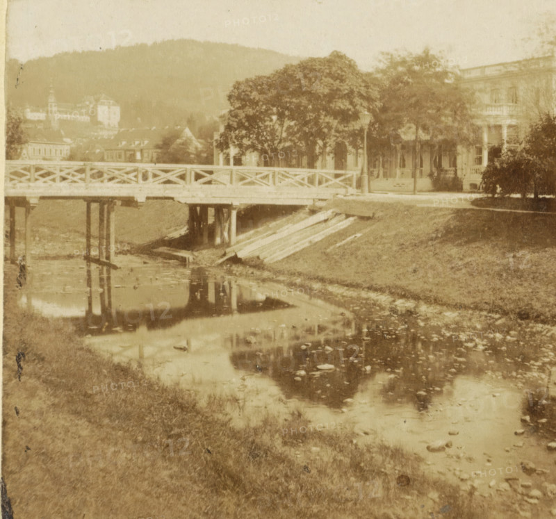 Villa Stephanie in Baden-Baden, Germany, Le Villa Stephanie, a Baden-Baden, Bords du Rhin, stereograph, inscription: 'ue, 80', number: 'No 34', Baden-Baden, 1861 - 1870, photographer, Baden-Württemberg, pasteboard, paper, albumen print, height 87 mm x width 172 mm, villa, prospect of village, silhouette of village, bridge in village across river, canal, A faded sepia scene showing a wooden bridge with a crisscross railing spanning a narrow body of water, the bridge supported by visible posts and reflected in the water below, a grassy bank leading up to the bridge with wooden planks or beams stacked near the edge, a row of trees and a lamppost near the far end of the bridge partially obscuring a large multi-story building with porches and balconies, a distant hillside and additional buildings faintly visible beyond the trees, and ripples and scattered debris on the water surface reflecting parts of the bridge and surroundings.