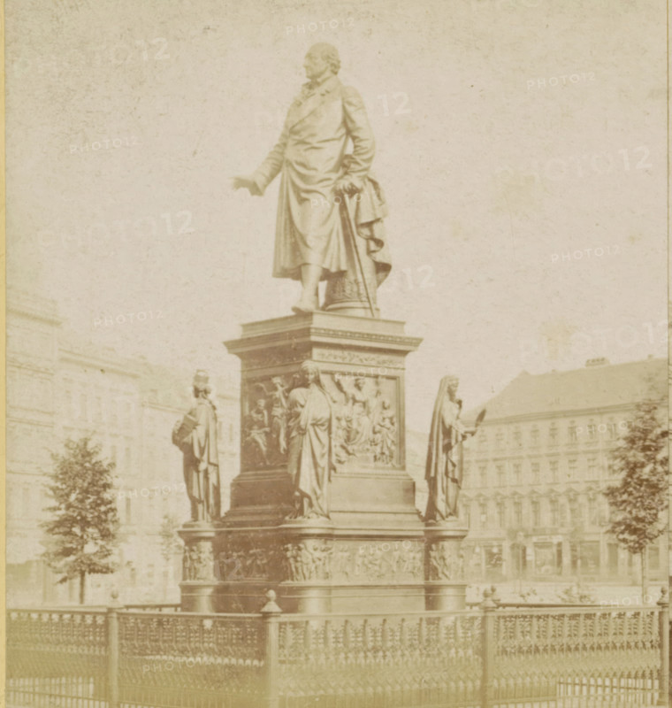 Das Stein-Denkmal, Statue of Baron van Stein in Berlin, Berlin and Environment, stereograph, photographer: Sophus Williams, Berlin, after sculpture by Hermann Schievelbein, 1877, Hermann Schievelbein, after sculpture by, pasteboard, paper, albumen print, height 86 mm x width 176 mm, Person, Heinrich Friedrich Karl vom und zum Stein, monument, statue, Dönhoffplatz, A tall stone statue of a robed man stands on a multi-tiered ornate pedestal surrounded by additional robed figures and sculpted relief panels, with a wrought iron fence in the foreground and large classical buildings and trees visible in the background.