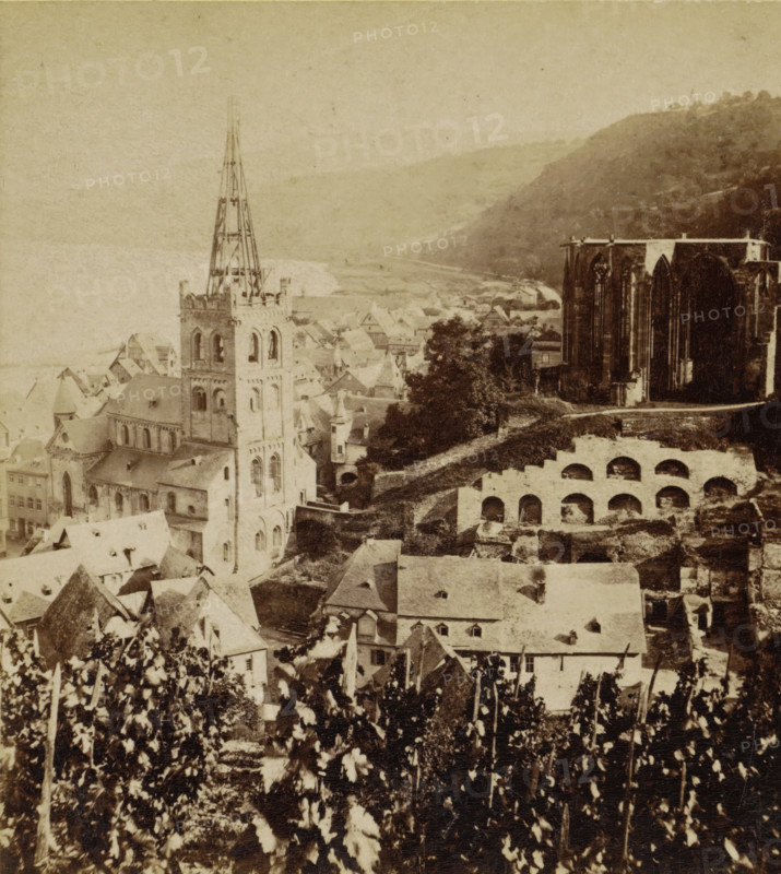 Bacharach. Panorama mit der Werner-Kapelle, Bacharach and the ruins of the Werner Chapel, Germany, Die Rheinlande, stereograph, number: '132.', photographer: Sophus Williams, Bacharach, 1877, Rhineland-Palatinate, pasteboard, paper, albumen print, albumendruk, height 86 mm x width 176 mm, city-view in general, 'veduta', dale, valley, ruin of church, monastery, etc. - QQ - small church, chapel, Wernerkapel, A sepia-toned aerial view of an old town with clustered rooftops and narrow streets, dominated by a tall church tower with an exposed spire structure and arched openings, adjacent to large Gothic ruins with high pointed window openings, terraced stone arches on a sloping hillside, scattered trees and foliage in the foreground, and distant hills fading into the pale sky.