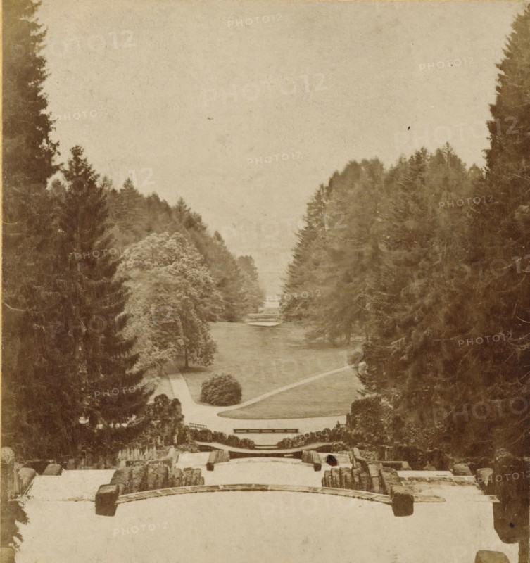 Wilhelmshöhe. Aussicht von den Cascaden, View from the large waterfalls in Wilhelmshöhe, Kassel, Cassel und Umgegend, stereograph, number: '10.', photographer: Sophus Williams, Berlin, 1876, Hessen, pasteboard, paper, albumen print, albumendruk, height 86 mm x width 177 mm, public gardens, park, garden cascade, Wilhelmshöhe, A sepia-toned photograph showing a formal landscape viewed from an elevated stone terrace with carved balustrades and steps in the foreground, a long central lawn or waterway flanked by paths and trimmed shrubs, rows of tall evergreen trees forming dense borders on both sides that taper toward the background, layered terraces and small bridges visible along the central axis, and a pale sky occupying the upper portion of the image.