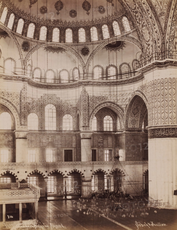 Interior of the Sultan Ahmet Mosque in Istanbul, Intérieur de la Mosquée Ahmed, photograph, number: '300', photographer: Sébah & Joaillier, Istanbul, 1888 - 1900, paper, albumen print, height 268 mm x width 208 mm, temple, shrine, Islam, Mohammedanism, Sultan Ahmet Mosque, The image shows an interior space with a large ornate dome and multiple tiers of arched windows and balconies. Intricate floral and geometric patterns cover the walls, arches, and columns, with decorative medallions and ornate borders visible along curved surfaces. Tall slender columns rise to support arches adorned with detailed motifs, and a series of hanging chains and light fixtures descend from the ceiling toward a central chandelier arrangement near the floor. The lower wall surfaces display patterned tilework and rows of arched doorways or niches, while the floor appears polished and reflective. A partial elevated wooden structure with balustrades is visible in the foreground, and a large plain structural pill...