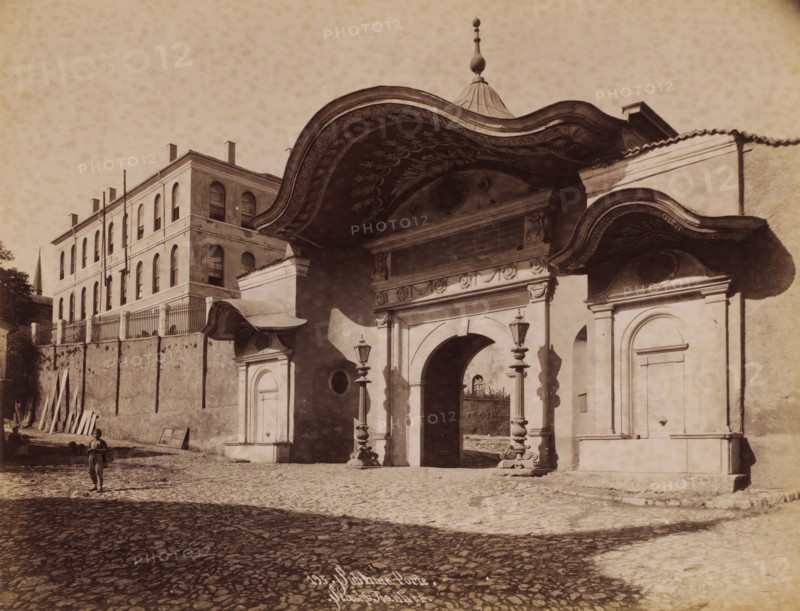 Sublime Porte in Istanbul, Sublime Porte, photograph, number: '195.', photographer: Sébah & Joaillier, Istanbul, 1888 - 1900, paper, albumen print, albumendruk, height 208 mm x width 268 mm, gate, entrance, A sepia-toned street scene showing a large ornate arched gateway with carved decoration and a curved, painted canopy above, flanked by smaller doorways and ornate lamp posts on stone pedestals; a multi-story building with arched windows and a fenced parapet sits behind a high wall to the left, with wooden planks leaning against the wall and a narrow spire visible in the distance; a cobblestone street fills the foreground where a solitary child stands wearing simple clothing and holding an object, casting a shadow on the ground.