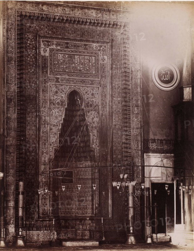 Interior of a mosque, possibly in Istanbul, photograph, photographer: attributed to Sébah & Joaillier, Istanbul, 1888 - 1900, Attributed to, paper, albumen print, albumendruk, height 270 mm x width 210 mm, temple, shrine, Islam, Mohammedanism, A sepia-toned photograph showing a section of a richly decorated interior wall with an ornate recessed niche framed by intricate geometric and floral patterns, scalloped arch detailing, and layered rectangular panels. The wall surface is covered in dense repeated motifs and borders, with vertical columns on either side that have carved or painted decorative bands. A large round medallion with concentric rings and an emblematic mark is visible on the right above a plain wall area. Hanging lamps and chandeliers suspended by thin wires or cords appear across the foreground, and tall slender columns with bases and capitals stand near a recessed doorway on the right. The lower portion shows a raised platform or step and detailed friezes along the base...