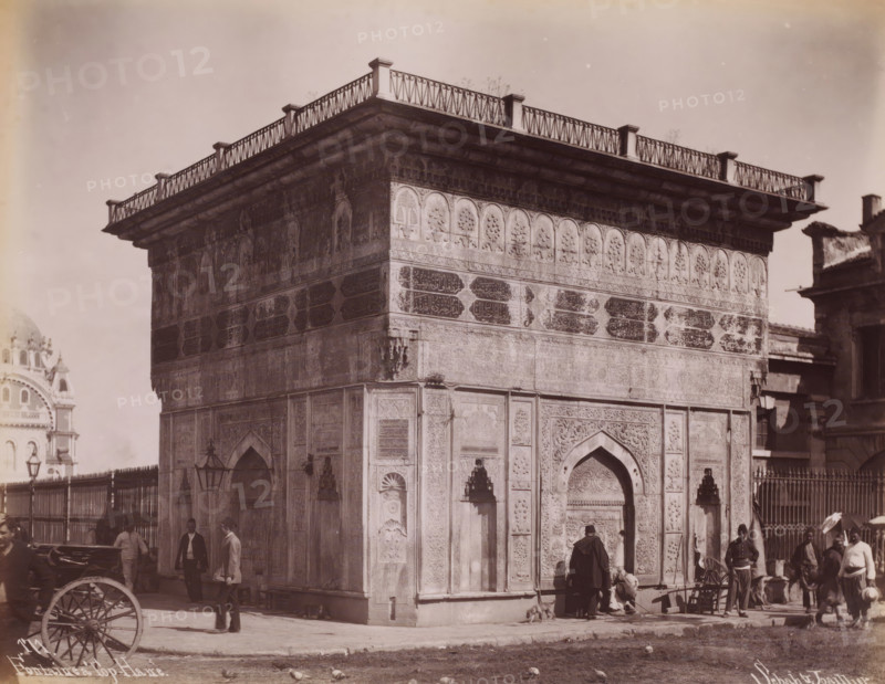 Fontaine a Top-Hane, Tophane fountain in Istanbul, photograph, number: '171.', photographer: Sébah & Joaillier, Istanbul, 1888 - 1900, paper, albumen print, albumendruk, height 210 mm x width 270 mm, utilitarian, public pump or water fountain, Tophane fountain, A corner view of a large ornate stone building with carved arches and decorative panels, a projecting cornice topped by a low railing with vertical posts and diagonal crosswork, and patterned friezes below the cornice; people stand and walk near the base of the building, some wearing long coats and hats and one person seated on the ground beside a small dog, a parked cart wheel is visible in the foreground, a metal fence and lamp posts run along one side, and portions of other buildings appear at the edges of the image.