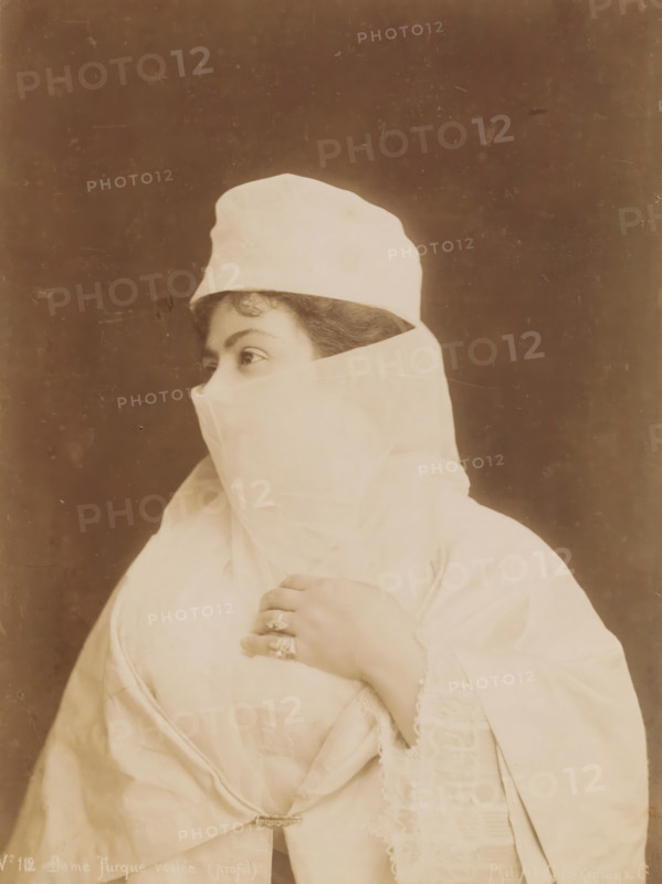 Portrait of an unknown veiled Turkish woman, Dame Turque voilée (profil), photograph, number: 'No 112', photographer: G. Lekegian & Co., Middle East, 1887 - c. 1900, paper, albumen print, height 275 mm x width 213 mm, adult woman, anonymous historical person depicted, A sepia-toned portrait of a person turned in profile wearing a light-colored head covering and veil that conceals the lower face, revealing only the eyes and a curl of hair at the forehead; the person’s hand is raised to the chest, showing multiple rings on the fingers and a bracelet at the wrist, and the clothing includes layered fabric with lace trim and a decorative clasp near the waist against a plain, dark background.
