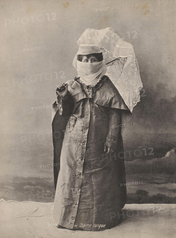 Portrait of an unknown veiled Turkish woman, Turkish lady, photomechanical print, number: '99.', Middle East, cliché maker: anonymous, 1870 - 1930, photographer, paper, collotype, photogravure, height 236 mm x width 180 mm, adult woman, anonymous historical person portrayed, A full-length sepia-toned photograph of a person standing and facing the camera, wearing a long, buttoned outer garment with a shiny, textured surface and a contrasting inner layer; the upper garment has a decorative collar and ruffled trim, and one sleeve shows a cuff. The person's face is mostly covered by a light-colored cloth wrapped around the lower face and head, leaving only the eyes visible under a small brimmed hat. The person holds an open lace-patterned parasol over a shoulder with one hand, while the other hand is raised near chest level holding a small object. The background is a plain, softly shaded backdrop and the floor beneath the figure shows a draped cloth.