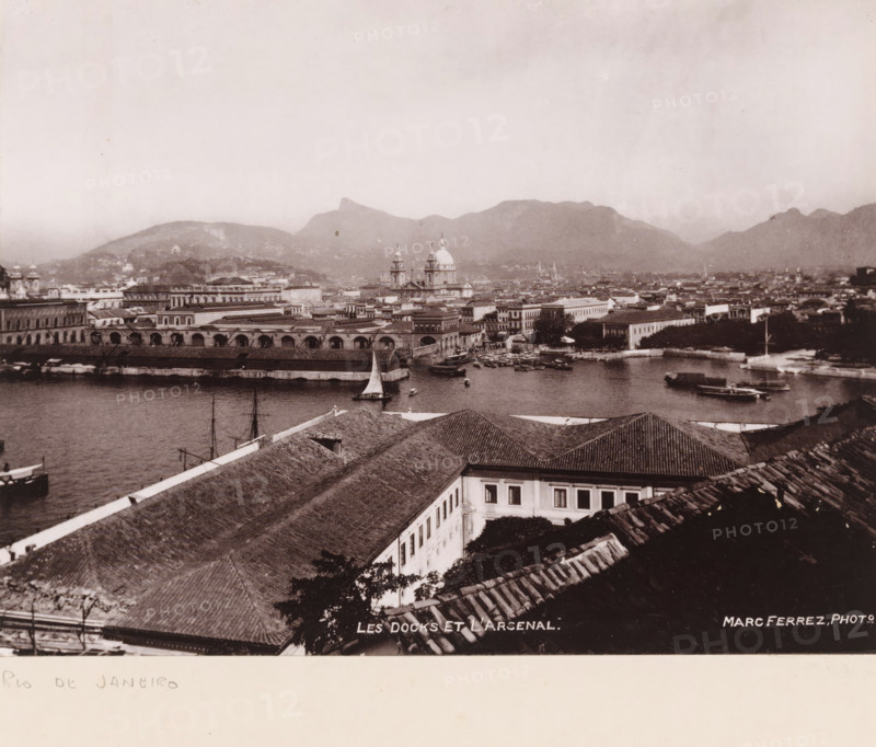 View of the port and the arsenal of Rio de Janeiro, Les docks et l'arsenal, photomechanical print, address: 'MARC FERREZ, RIO DE JANEIRO, 88, RUA SÃO JOSÉ', photographer: Marc Ferrez, Rio de Janeiro (city), cliché maker: anonymous, 1890 - 1910, Marc Ferrez, photographer, cliché maker, Sudeste, pasteboard, paper, collotype, photoprint, height 150 mm x width 202 mm, height 259 mm x width 332 mm, harbour, ammunition store, arsenal, A sepia-toned aerial view of a coastal city showing a waterfront with several boats and sailboats moored in calm water, a long dockside building with arched openings along the water, and a cluster of low-rise buildings with tiled roofs in the foreground. Beyond the waterfront are more multi-story buildings, a prominent church with domes and towers rising above the surrounding structures, and additional architectural details across the harbor. In the background a range of mountains forms a silhouette beneath a pale sky.