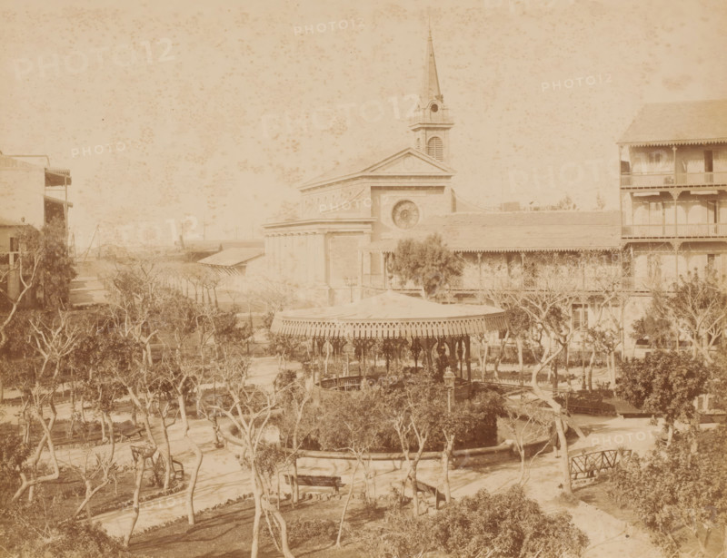 View of the Place de Lesseps in Port Said, Place de Lesseps, Port Saïd, photograph, inscription: 'Place de Lesseps Port Said, photo Zangaki, Port Said', photographer: Zangaki, C. & G., Port Said, 1870 - 1910, North Africa, pasteboard, paper, albumen print, height 219 mm x width 276 mm, height 347 mm x width 453 mm, square, circus, church (exterior), merry-go-round, roundabout, chair o'plane, A faded sepia photograph of a park scene with a central gazebo featuring a decorative roof and ornate pillars surrounded by trees and winding paths, benches placed near the paths, and a circular low wall around a planted area; behind the gazebo is a large classical building with a triangular pediment, a circular window, and a tall steepled tower, flanked on the right by a multi-story building with balconies and on the left by another building with exposed verandas, all seen under a speckled sky.