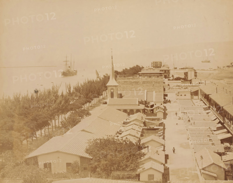 View of Port Tewfik, near the Suez Canal, Port Tewfik Suez, photograph, number: 'No. 138', inscription: 'Port Tewfik Suez, foto Zangaki', photographer: Zangaki, C. & G., Port Tewfik, 1870 - 1910, North Africa, Suez, pasteboard, paper, albumen print, albumendruk, height 219 mm x width 275 mm, height 348 mm x width 453 mm, city-view in general, 'veduta', canal, Suez Canal, A sepia-toned coastal scene showing a row of tiled-roof buildings and houses extending along a street toward the waterfront, with a church tower and steeple rising among the rooftops, trees lining a promenade beside the water, several sailing vessels and ships anchored or sailing on a calm sea, a few small figures walking in the street, and low-lying structures and vegetation along the shoreline.